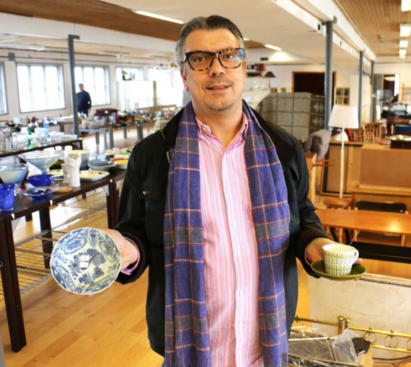 A man with medium-length wavy hair, wearing a teal work jacket, stands looking directly at the camera with a serious expression. The background features blurred white containers, suggesting a workplace environment.