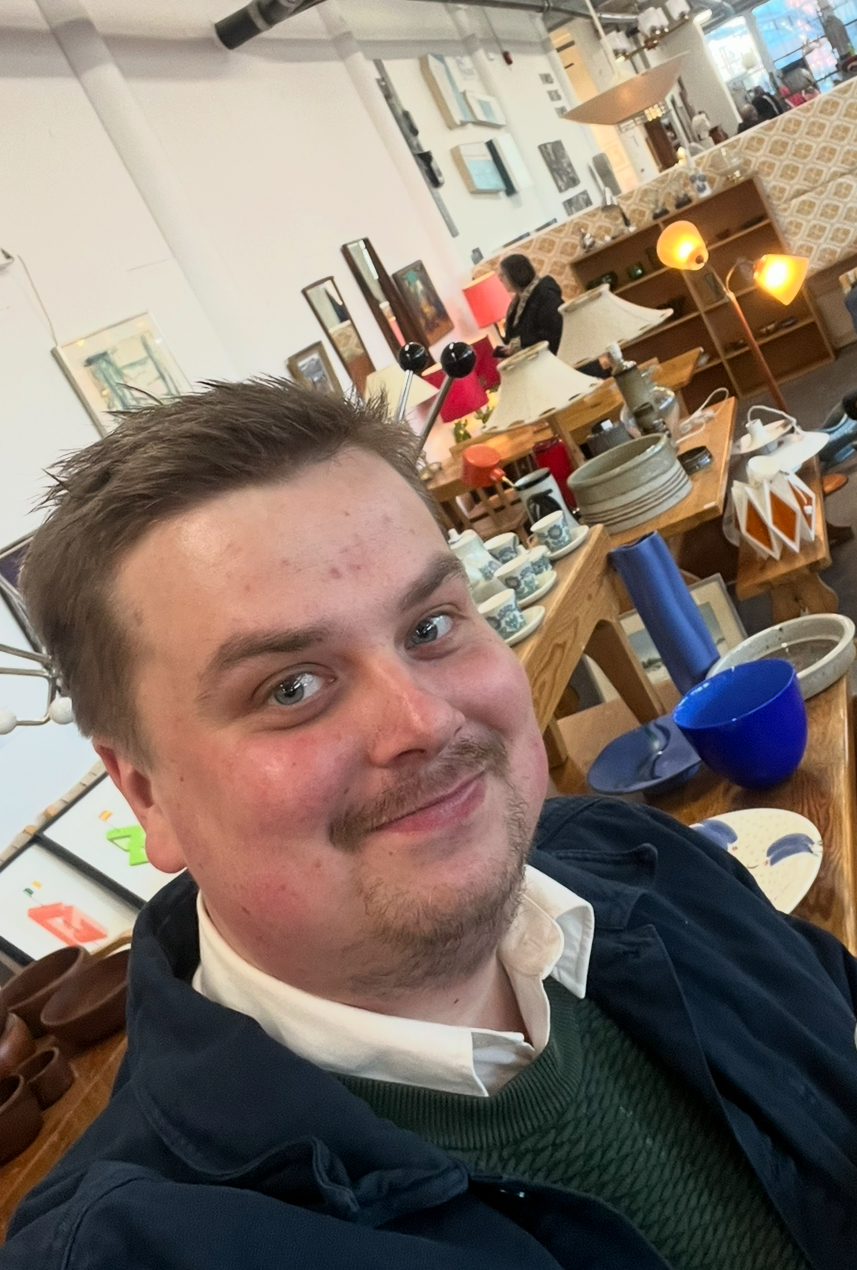 A woman with long, wavy hair, wearing a white button-up shirt, smiles confidently against a light gray background.