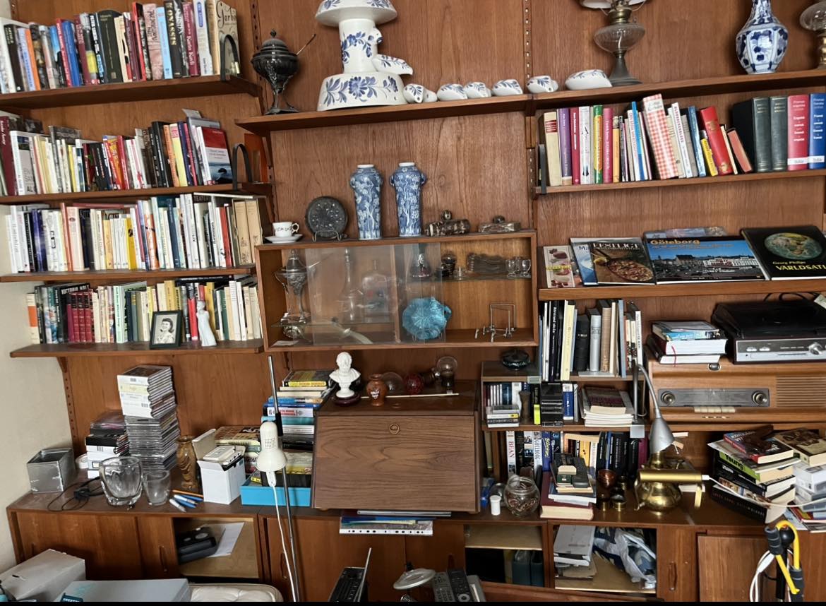 A young woman with medium-length wavy hair wearing a plain white t-shirt, smiling warmly in a bright, modern interior with bookshelves in the background.